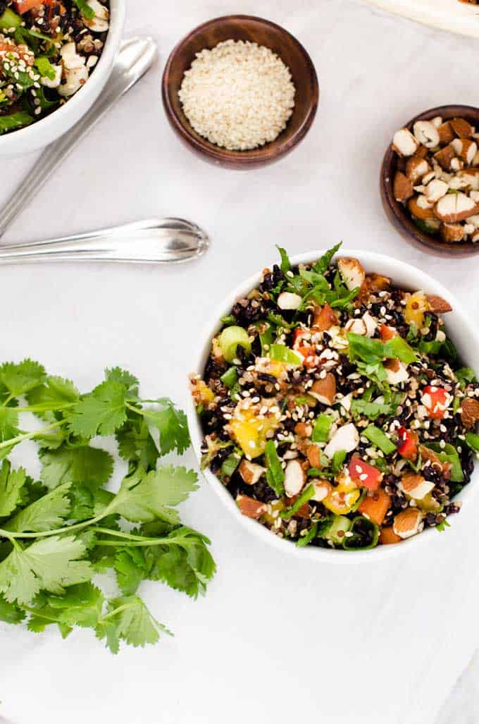 Overhead photo of Thai Quinoa Salad in a white bowl against a white background with small bowls of sesame seeds and almonds above and a bunch of cilantro below.