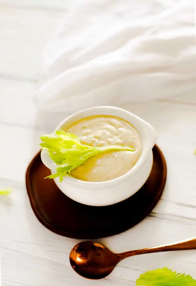 Photo of Cream of Celery Soup garnished with celery leaves in a white bowl sitting on a small dark wooden plate.