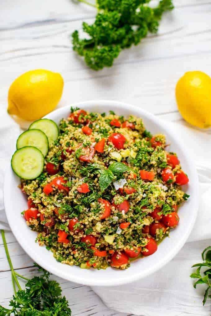 Overhead photo of a shallow bowl of Quinoa Tabbouleh garnished with mint with mint, parsley and lemons scattered around.