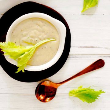 Square photo of Cream of Celery Soup garnished with celery leaves in a white bowl sitting on a small dark wooden plate.