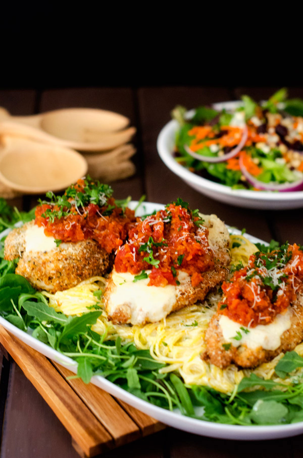 A platter with three pieces of chicken parmesan over pasta surrounded by baby spinach.