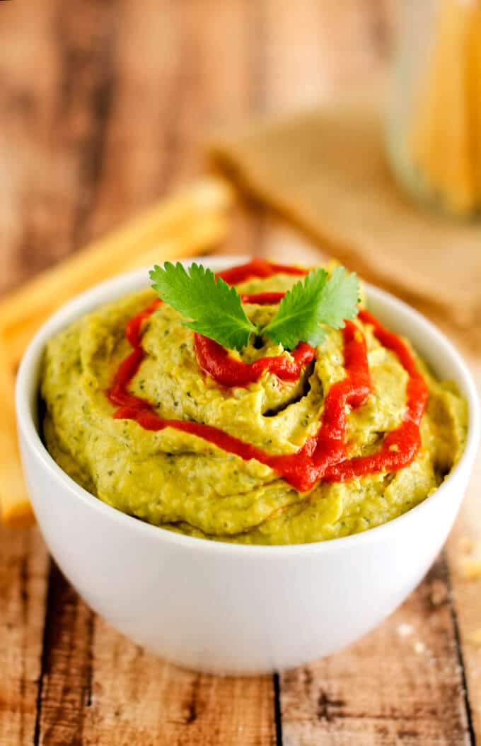 Close up photo of Avocado Hummus with Chickpeas in a small white bowl with breadsticks around it.