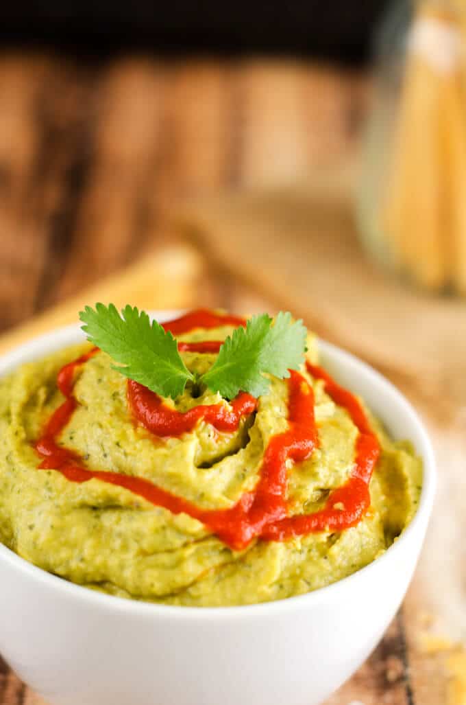 Close up shot of Avocado Hummus with Chickpeas in a small white bowl drizzled with Sriracha and garnished with cilantro.