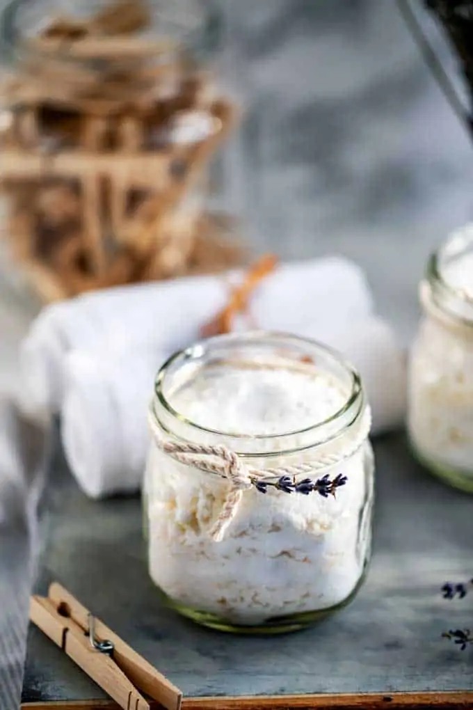 Photo of DIY Laundry Detergent in a small glass jar with a sprig of lavender tied around it and a jar of clothespins behind it.