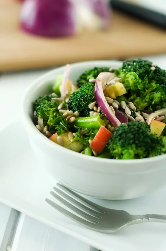 Side photo of a broccoli apple salad in a white bowl sitting on a white plate.