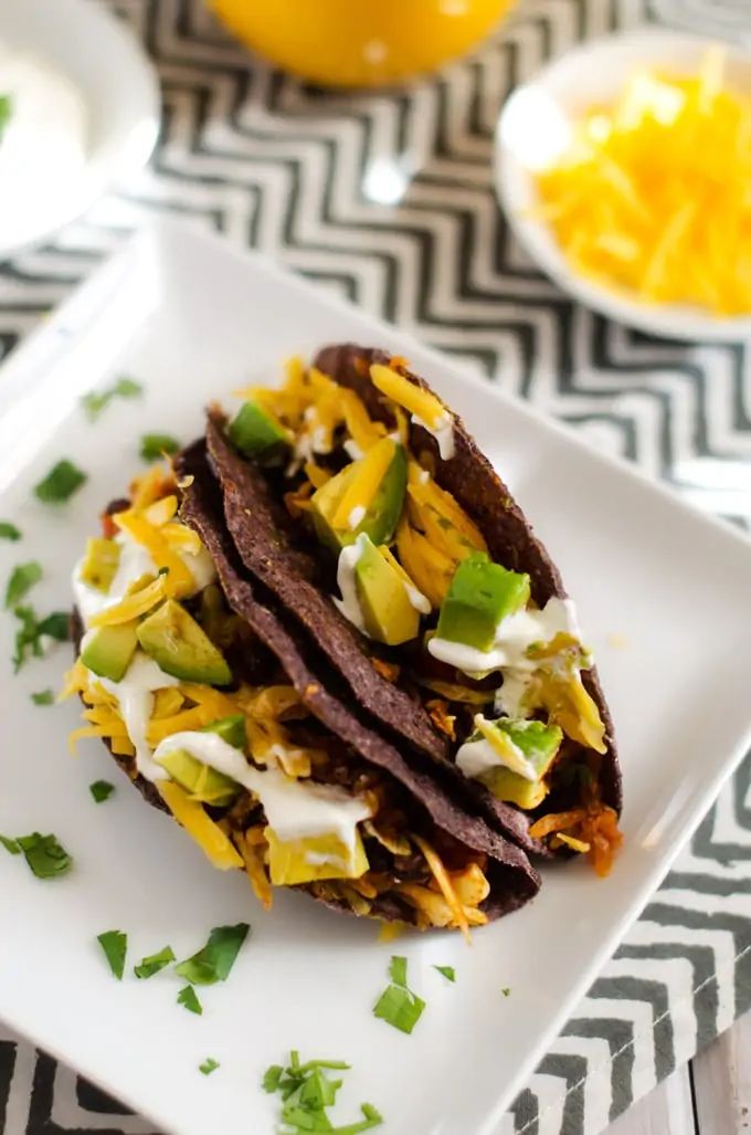 Two black bean tacos on a white plate with a small bowl of shredded cheese behind it.
