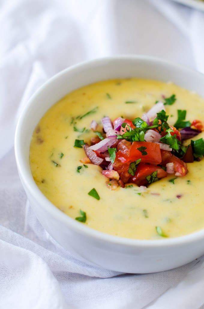 Homemade Queso Dip in a white bowl garnished with red onion, tomato, and cilantro.