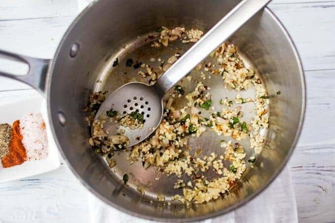 Onion and garlic being sauteed in a sauce pan with a spoon in it sitting on a white background.