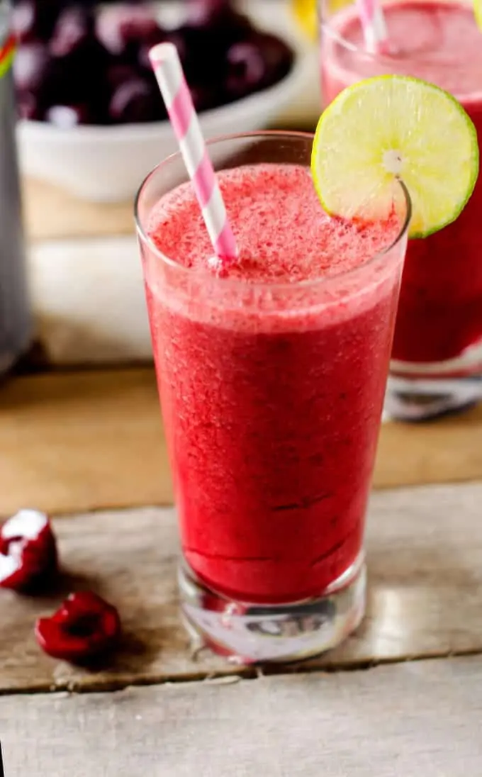 Photo of a Cherry Smoothie with a lime garnish sitting on a wooden board with two fresh cherries beside it and a bowl of cherries behind it.