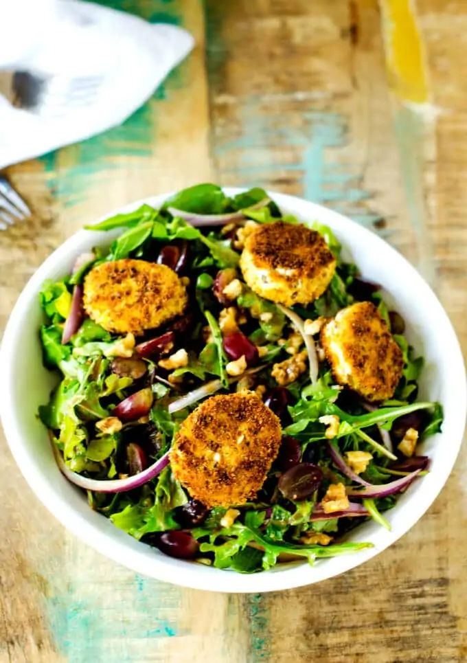 Overhead photo of a fried goat cheese salad on a white bowl against a rustic wooden background.