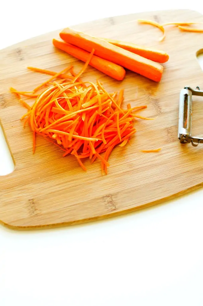 Photo of peeled carrots, a vegtable peeler, and shredded carrots on a cutting board.