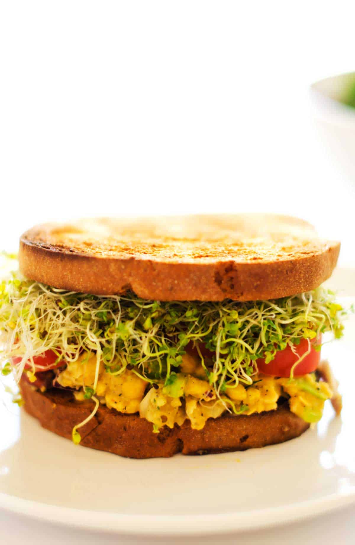 Close up side photo of a chickpea salad sandwich on a white plate agains a white background.
