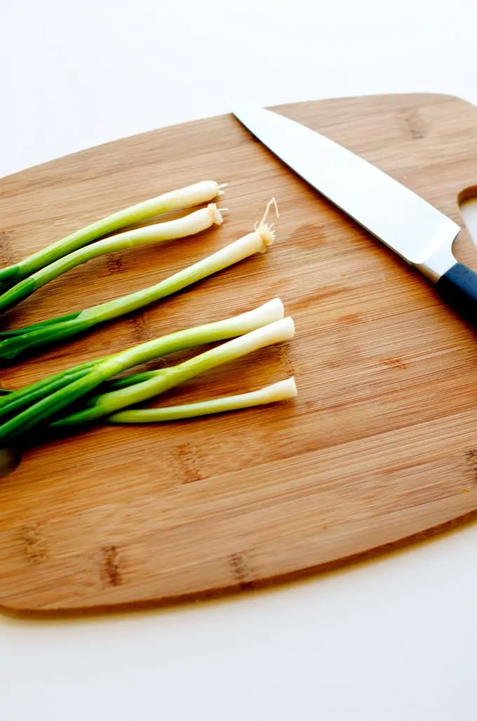 Photo of green onions and a knife on a cutting board agains a white background.