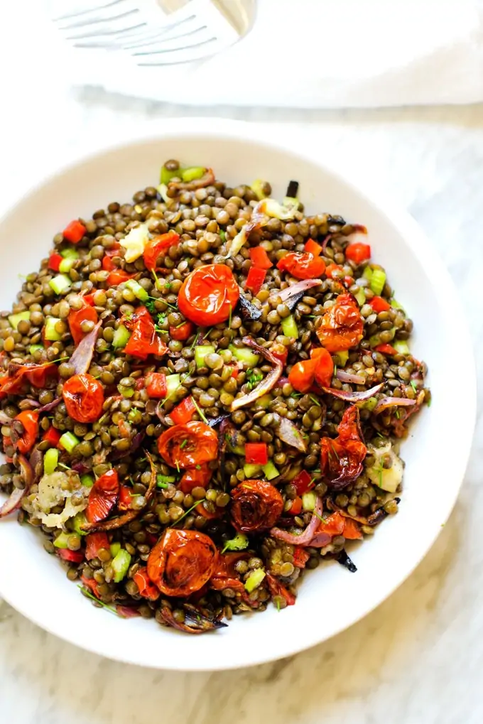Photo of a lentil salad with roasted tomatoes, onions, and garlic in a white serving dish sitting on a white marble background.