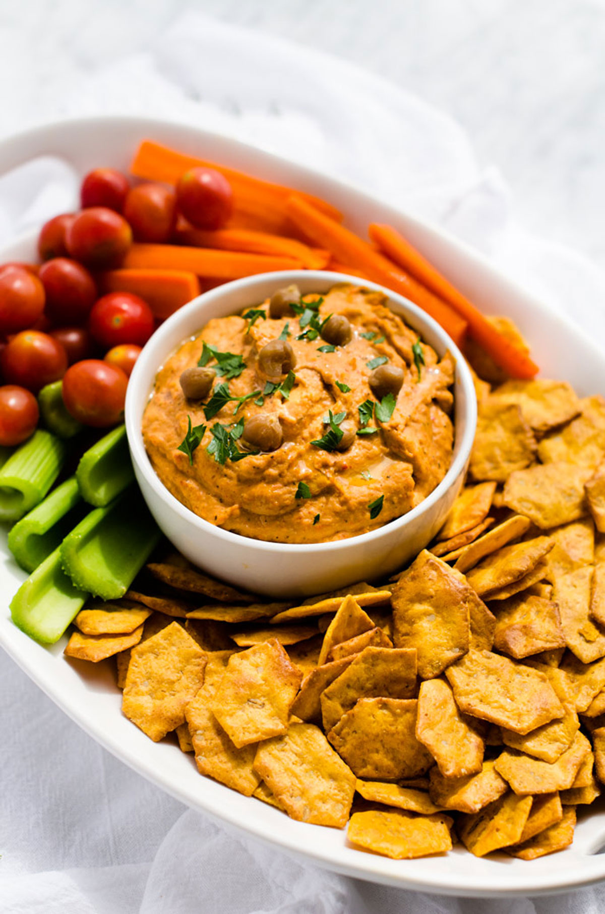Photo of a platter with red pepper hummus surrounded by crackers and fresh vegetables.