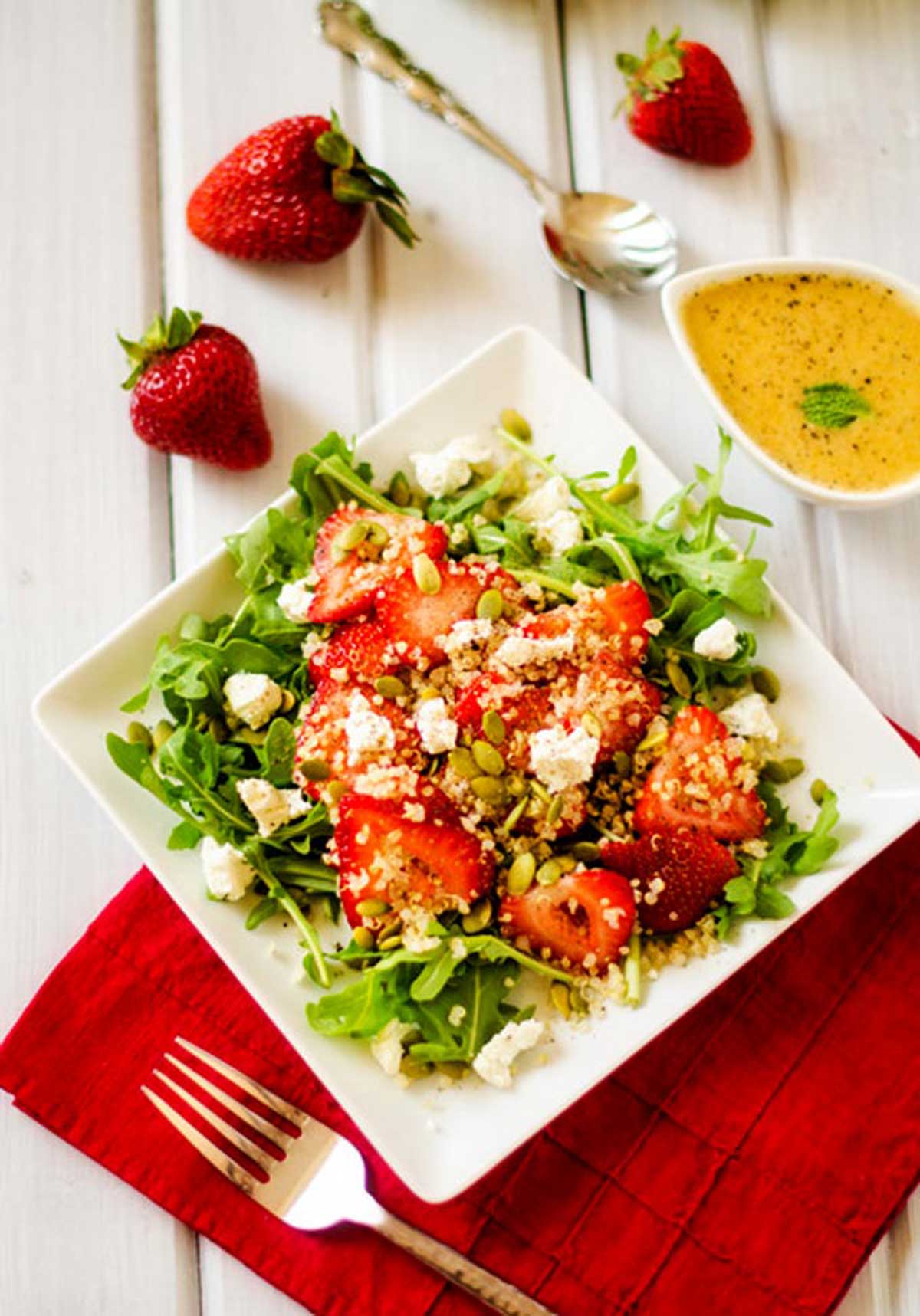 A white background with a strawberry quinoa salad on a square plate sitting on a red napkin.