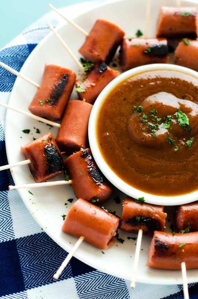 Photo of a honey mustard dip in a small white bowl surrounded by sausages on skewers.