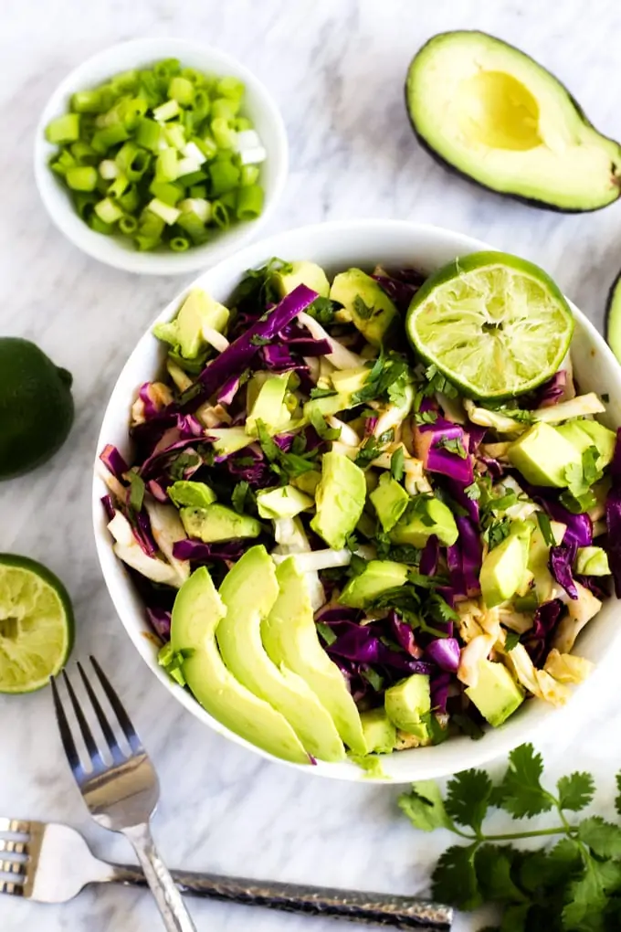 Overhead photo of a white bowl with Cilantro Lime Coleslaw in it surrounded by avocado, cilantro, limes, and green onions.