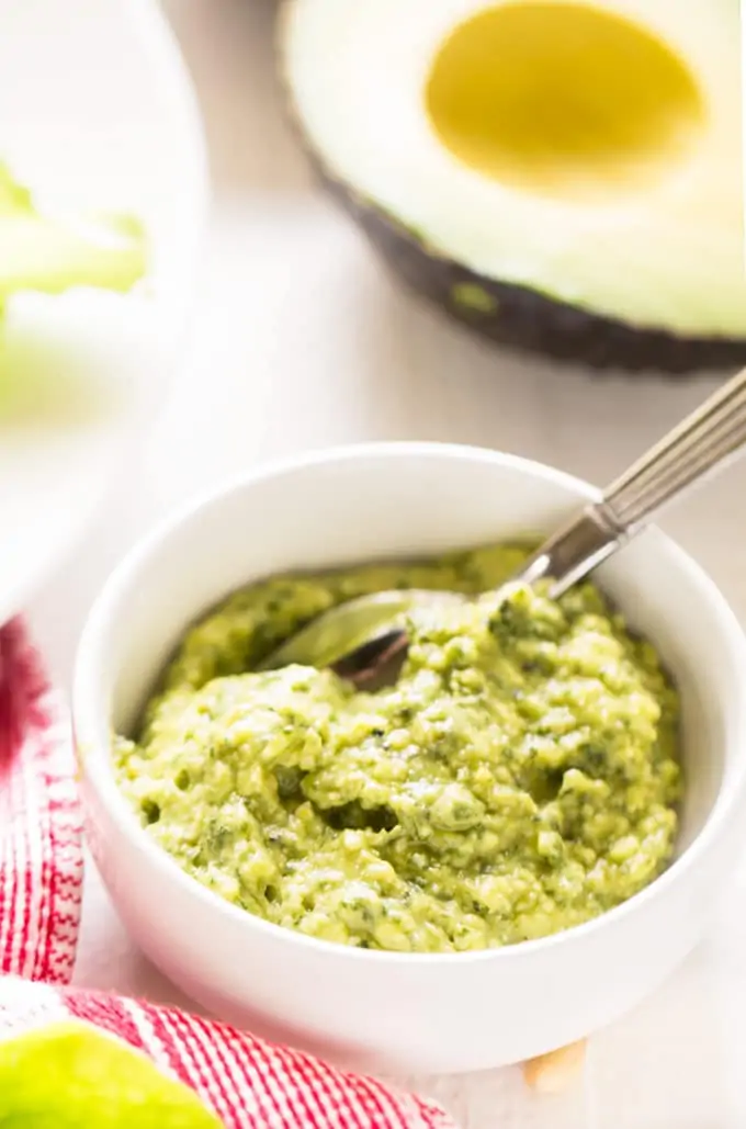 Photo of avocado pesto in a white bowl with a spoon in it and an avocado behind it.