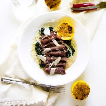 Square photo of a shallow white bowl with steak with spinach in it garnished with lemons and parmesan.