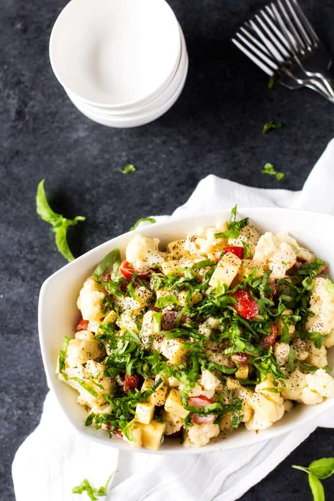 Overhead photo of Cauliflower Potato Salad in a large white bowl on a grey background with bowls to serve in and forks behind the salad.