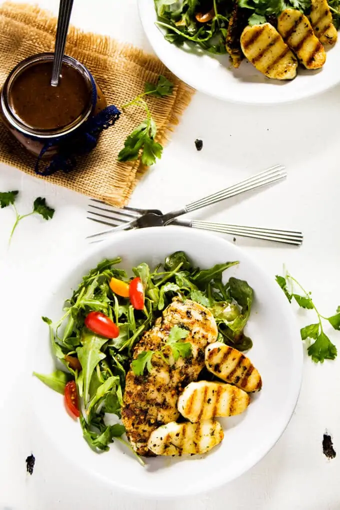 Overhead photo of a Chicken Halloumi Salad on a white plate with a jar of Balsamic Vinaigrette behind it on a white background. 