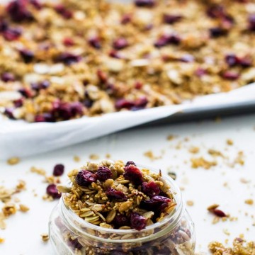 Photo of quinoa granola in a small glass jar on a white background with a sheet pan of quinoa granola behind it.