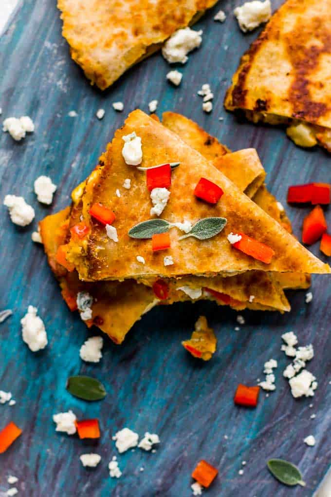 Butternut Squash Quesadillas on a blue background garnished with blue cheese, red pepper and small sage leaves.