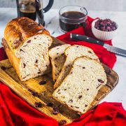 A loaf of cranberry bread, partially sliced, sits on a wooden cutting board with a red cloth underneath. Nearby are a knife, a bowl of cranberries, rich chocolate toffee pieces, a French press, and a cup of coffee.