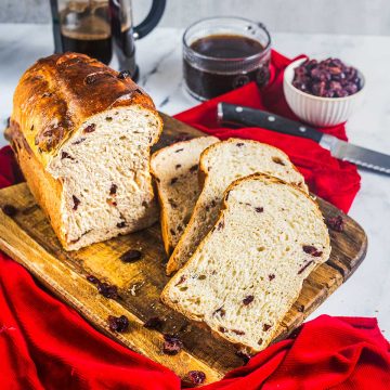 A loaf of cranberry bread, partially sliced, sits on a wooden cutting board with a red cloth underneath. Nearby are a knife, a bowl of cranberries, rich chocolate toffee pieces, a French press, and a cup of coffee.