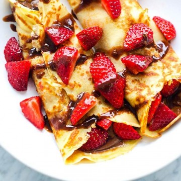 Overhead photo of gluten free crepes on a white plate with strawberries and chocolate hazelnut sauce on top.