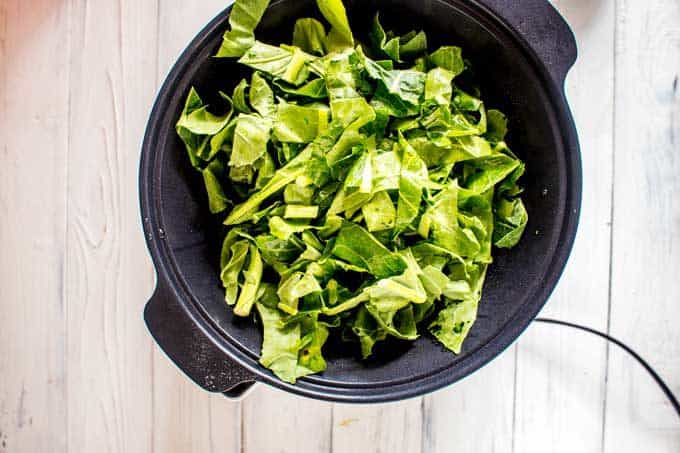 Photo of collard greens being added to a slow cooker sitting on a white background.