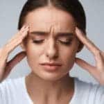 Photo of a woman in a white shirt holding both sides of her head with a tension headache.