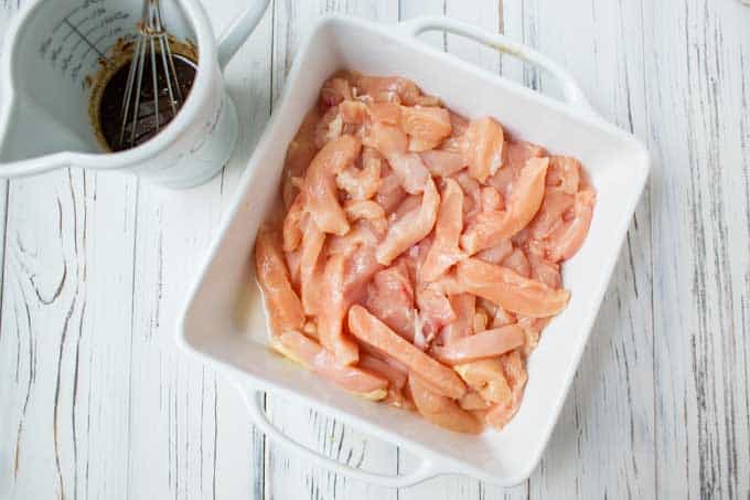 Chicken cut into strips in a white shallow dish with the marinade in a white liquid measuring cup with a whisk in it.