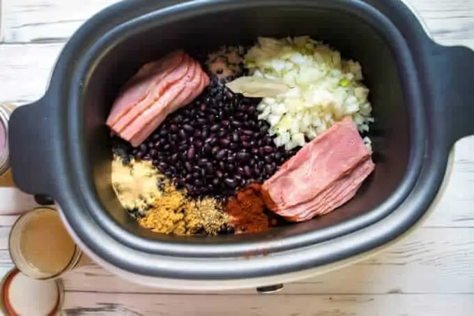 Photo of dried soaked black beans, chopped onion, bacon, and seasonings in a slow cooker ready to be cooked.