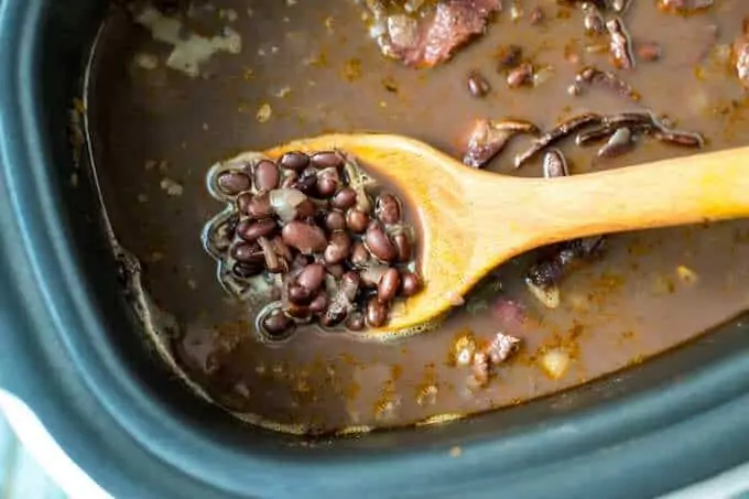Photo of a wooden spoon lifting out a spoonful of cooked black beans from a slow cooker.