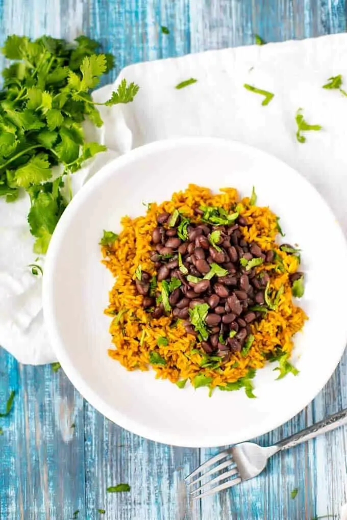 Photo of Slow Cooker Black Beans on a bed of turmeric rice on a white plate against a weathered blue background.  A bunch of parsley is above the plate with cut parsley spread around.