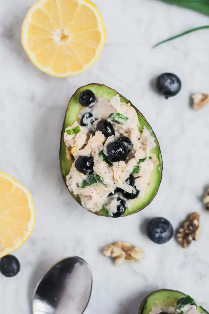 Aerial photo of an avocado stuffed with gluten-free blueberry chicken salad surrounded by blueberries, lemon wedge, walnuts, and green onions.