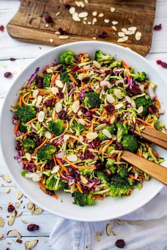 Close up photo of Broccoli Slaw Salad in a large white bowl with almonds and cranberries scattered on a white background.