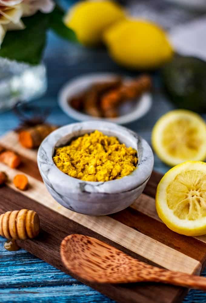 Photo of a prepared DIY Turmeric Face mask in a small bowl sitting on a wooden cutting board with fresh turmeric, lemons, and an avocado scattered around.