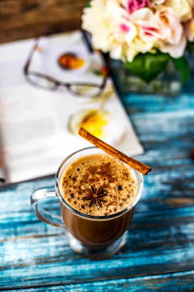 Photo of a Dirty Chai Latte in a glass mug garnished with a star anise and cinnamon with flowers, a magazine and reading glasses sitting behind it.