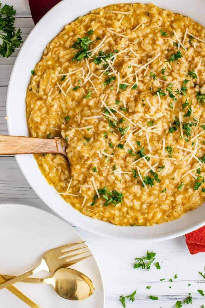 Overhead photo of Instant Pot Risotto in a large white serving bowl with a spoon in it. Plates with festive gold knives and spoons are sitting next to the bowl.
