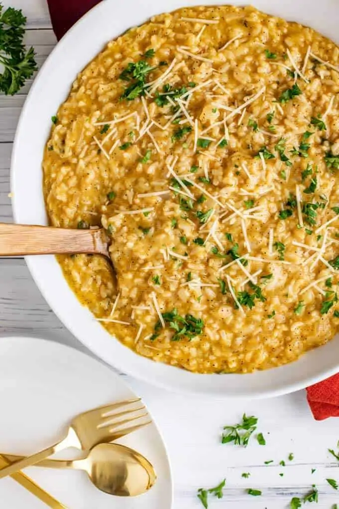 Overhead photo of Instant Pot Risotto in a large white serving bowl with a spoon in it. Plates with festive gold knives and spoons are sitting next to the bowl.