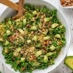 Overhead photo of Kale and Brussels Sprouts salad in a large white bowl with salad mixers in it and avocado, pecans and parsley sitting by it.