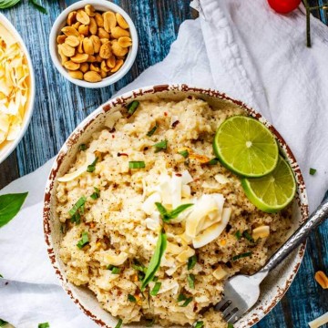 Overhead photo of Coconut Cauliflower Rice in a rustic white bowl garnished with Thai Basil, coconuts, and limes, sitting on a white napkin that rests on a blue table.
