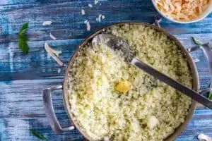 Photo of garlic being added to a skillet of cauli-rice that sits on a blue background.