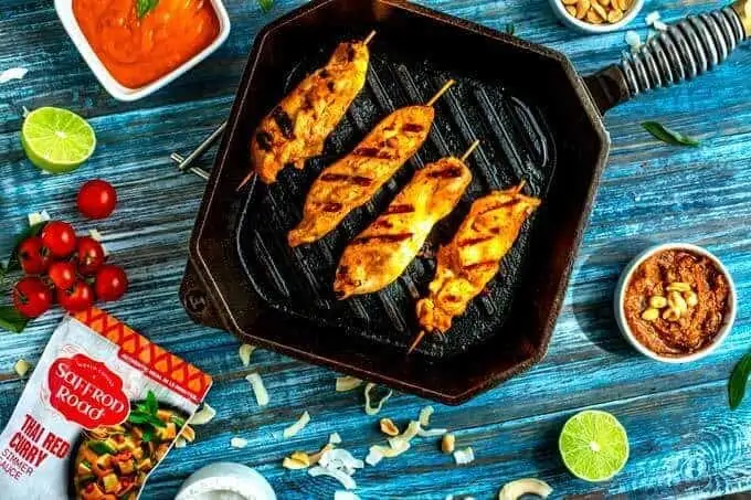 Photo of chicken strips on skewers in a cast iron skillet sitting on a blue background surrounded by sauce, pecans, coconut, tomato, and lime.