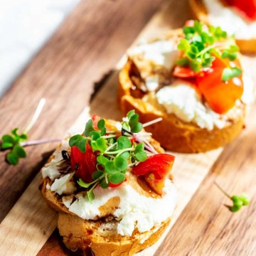 Close up photo of a small bread round topped with goat cheese, tomatoes, balsamic reduction and microgreens sitting on a wooden cutting board.