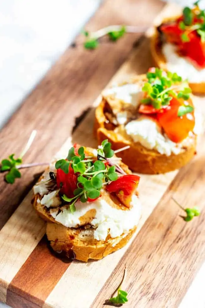 Close up photo of a small bread round topped with goat cheese, tomatoes, balsamic reduction and microgreens sitting on a wooden cutting board.