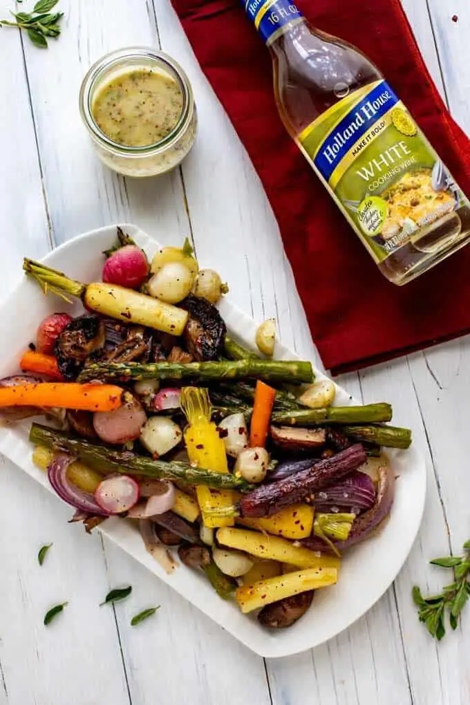 Overhead photo of Roasted Spring Vegetables on a white platter sitting on a wooden white background with thyme next to it and a bottle of Holland House White Cooking Wine.